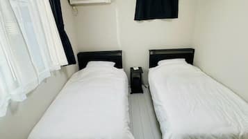 A simple room with two beds, a nightstand, an air conditioning unit, and a window with sheer curtains.