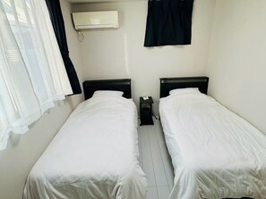 A simple room with two beds, a nightstand, an air conditioning unit, and a window with sheer curtains.
