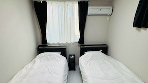 A room with two single beds, a window with curtains, an air conditioning unit, and a small table.