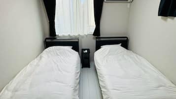 A room with two single beds, a window with curtains, an air conditioning unit, and a small table.