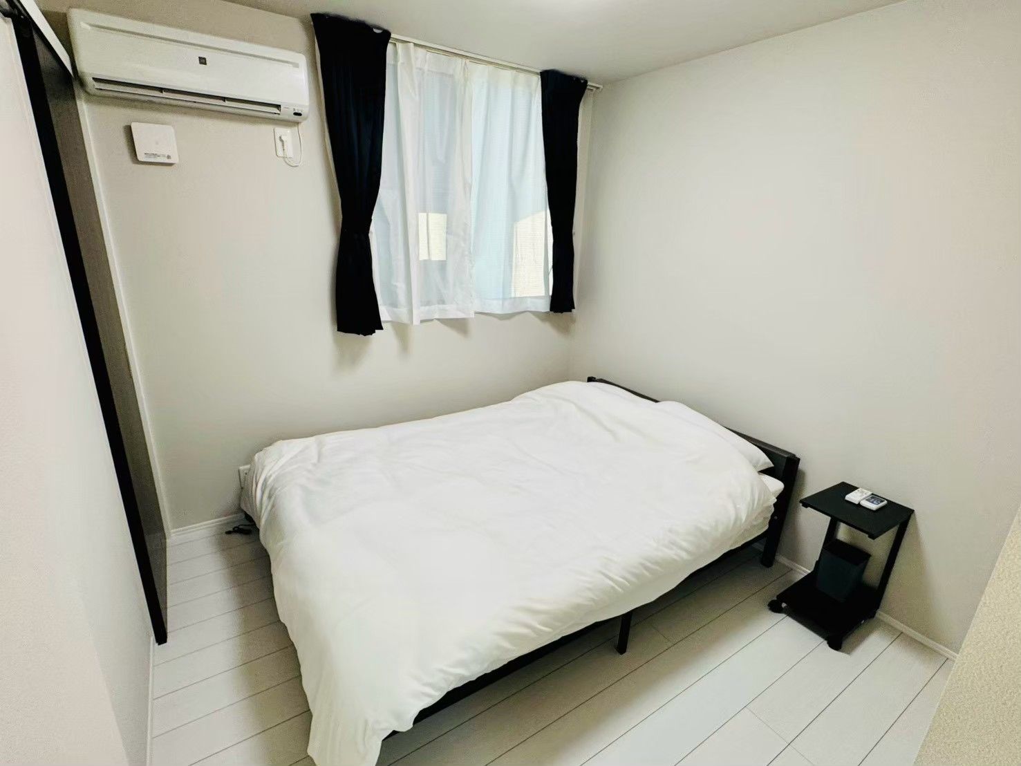 A small, single-bed room with a white bed, a black nightstand, a white air conditioning unit, and a window with black curtains.