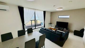 Un salon moderne doté d’un grand téléviseur, d’un canapé, d’une table à manger avec des chaises et offrant une vue sur l’extérieur grâce à une grande fenêtre.