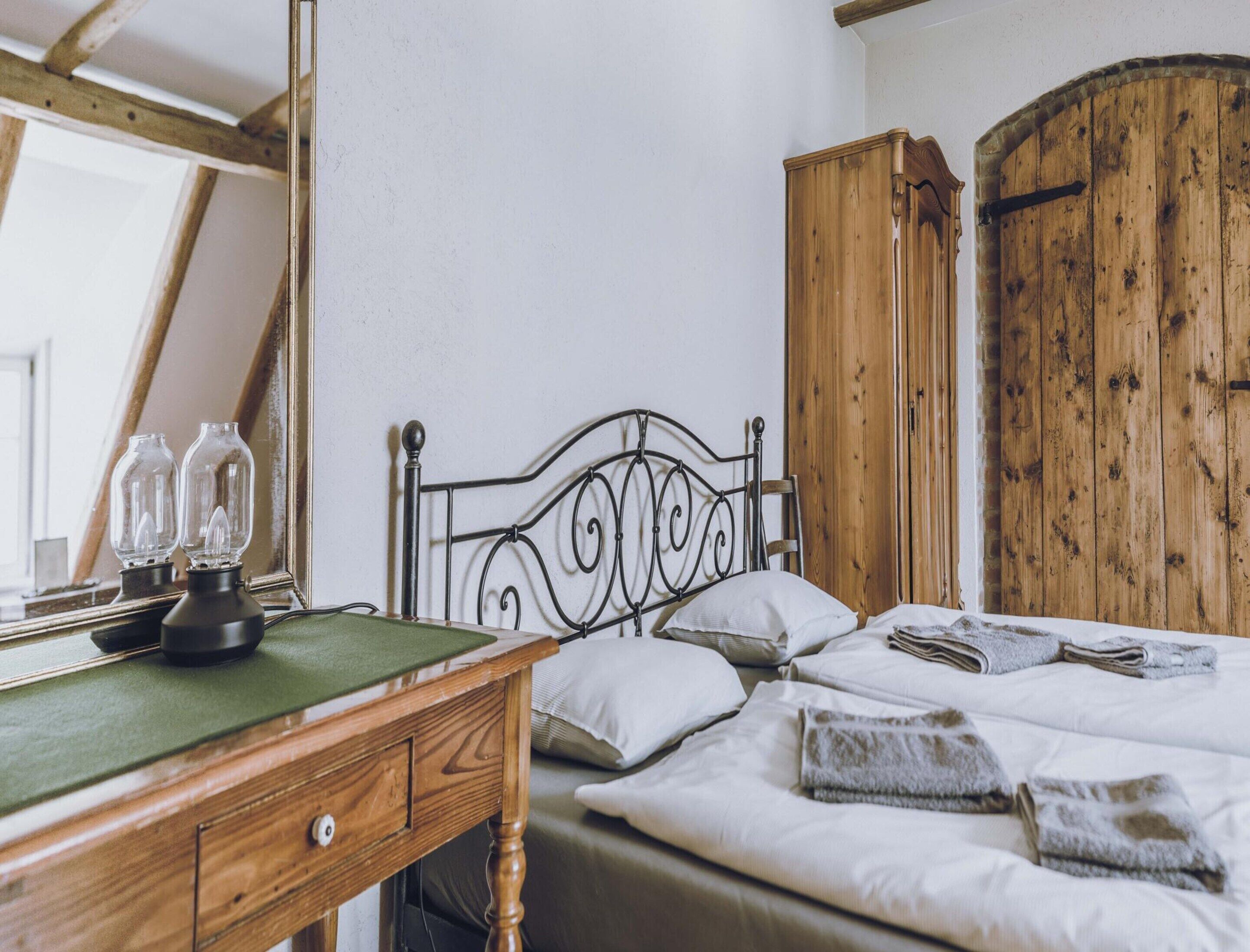 A bedroom with a wooden desk, a metal bed frame, a large wooden wardrobe, and a mirror.