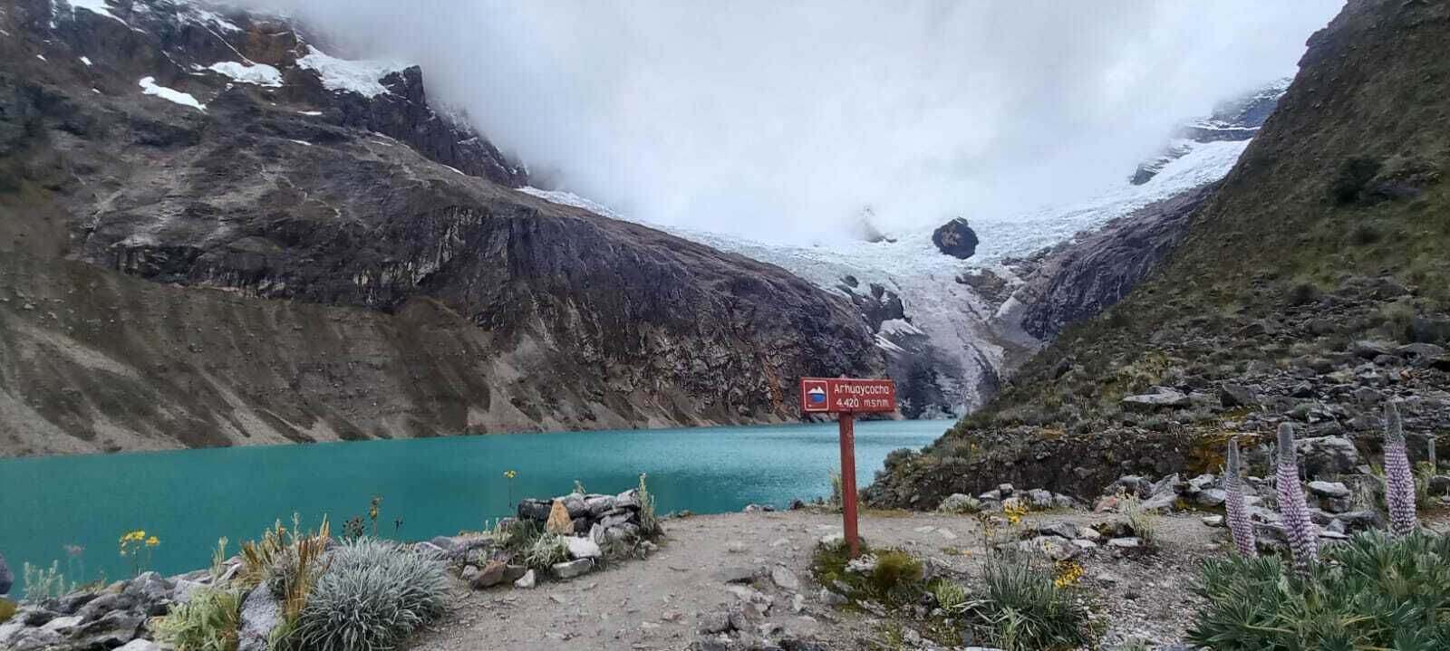 Paisagem montanhosa com um lago azul-turquesa, primeiro plano rochoso com vegetação escassa e um sinalizador indicando um local.