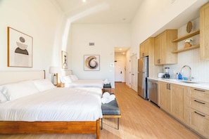 Habitación de hotel moderna con una cama grande, un banco, una kitchenette y un baño visible al fondo.