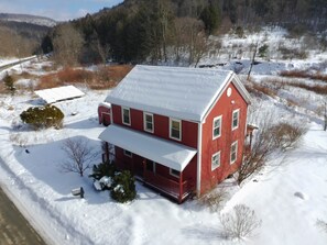 A red two-story house with a snow-covered roof, surrounded by a snowy landscape and a forested hill in the background. - Red Farmhouse By The Creek (Scott Township)