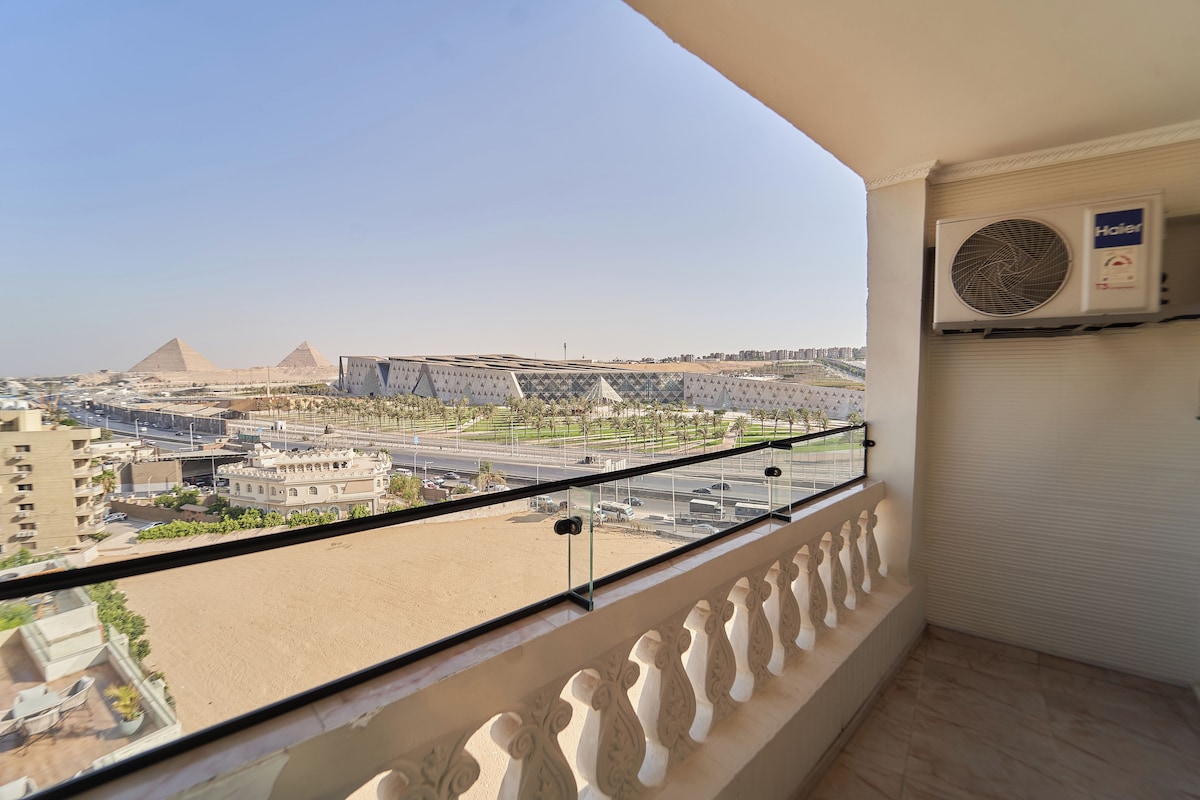 A balcony with a view of the pyramids, an air conditioning unit, and a modern building.