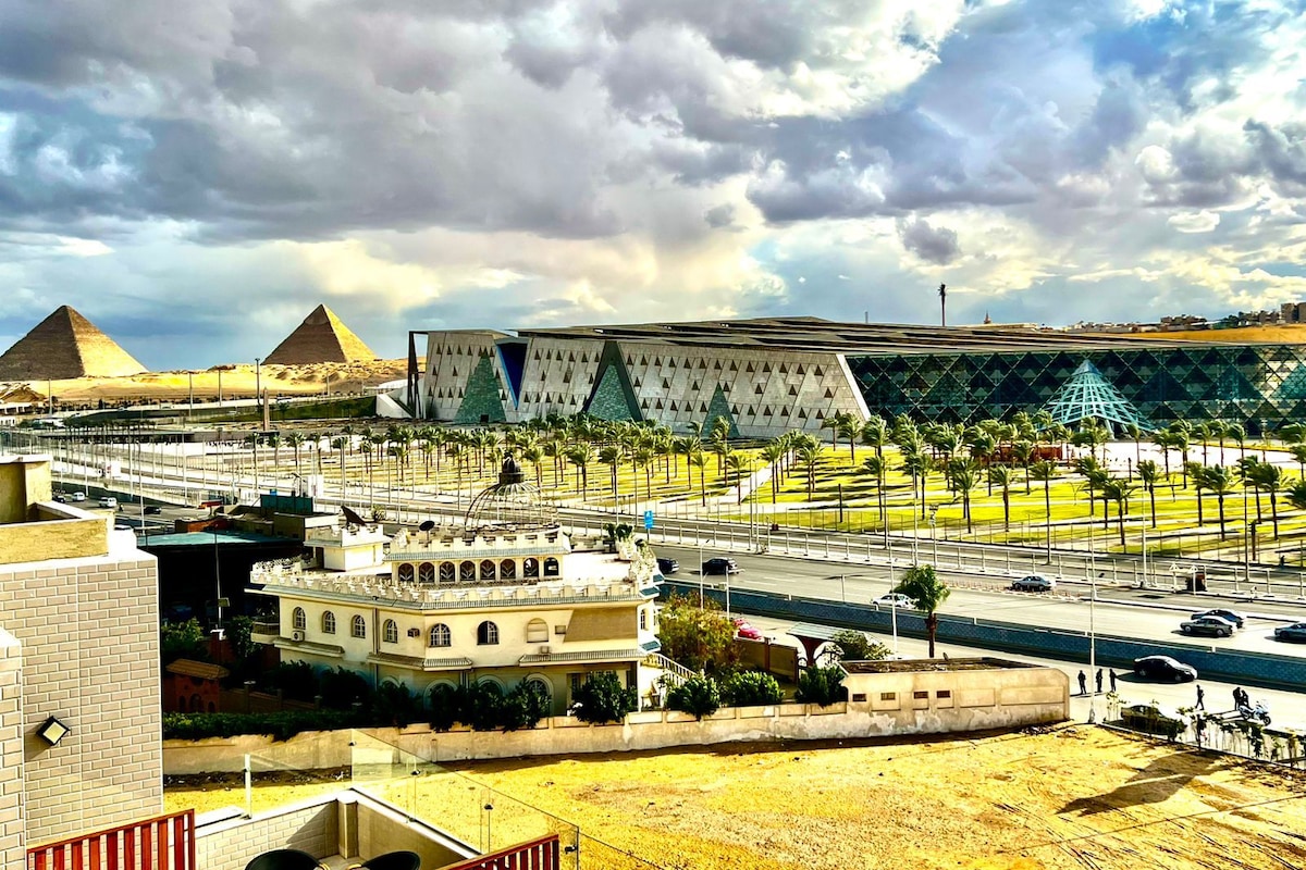 A modern building with a geometric facade, a large open plaza with palm trees, and pyramids in the background.