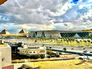 A modern building with a geometric facade, a large open plaza with palm trees, and pyramids in the background. - GEM View grand Boutique (Giza)