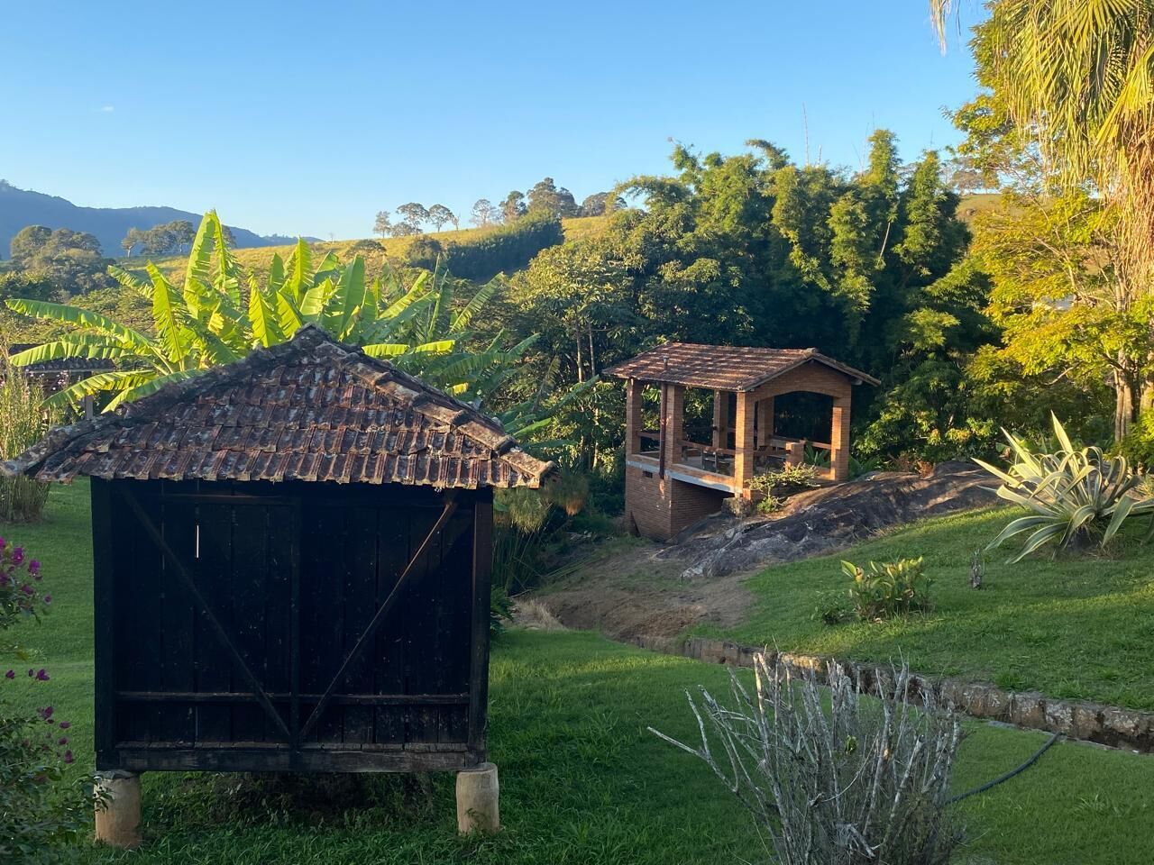 A rustic wooden structure with a tiled roof, surrounded by lush greenery and a hilly landscape.