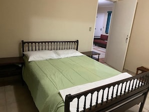 A single bed with a wooden frame and green bedding in a room with plain walls and a tiled floor.