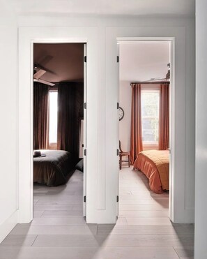 Un couloir mène à deux chambres dotées de planchers en bois. L’une d’elles est équipée d’un ventilateur de plafond, et l’autre possède un lit avec une tête de lit.