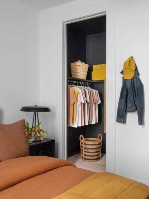 Un dormitorio con cama, mesita de noche, lámpara, armario con ropa colgada y una cesta.