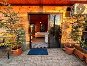 Una cabaña de madera con un vidrio en la puerta que da a un dormitorio con cama y mesita de noche. Hay plantas en macetas afuera y un felpudo azul en la entrada.