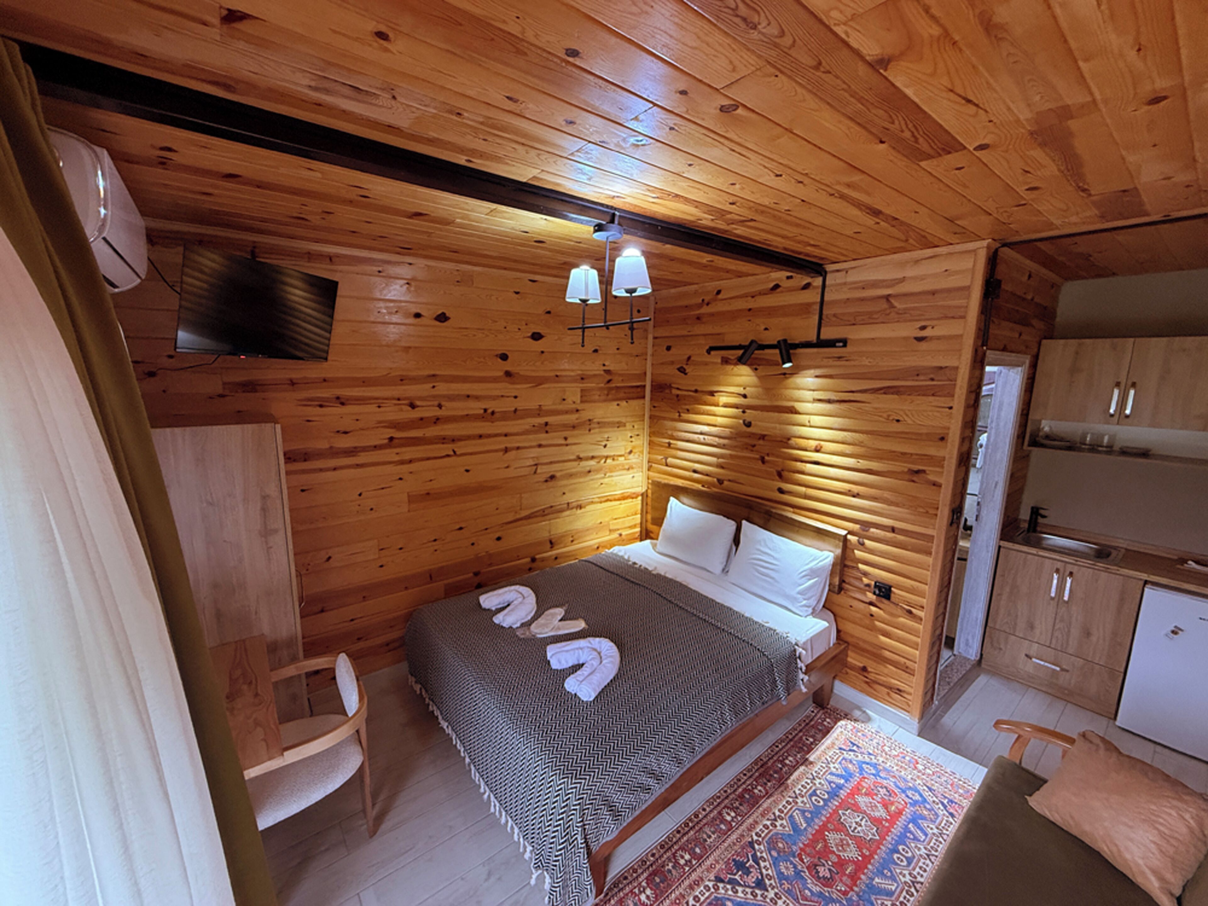 Interior de una cabaña de madera con cama, sillón, televisor y kitchenette.