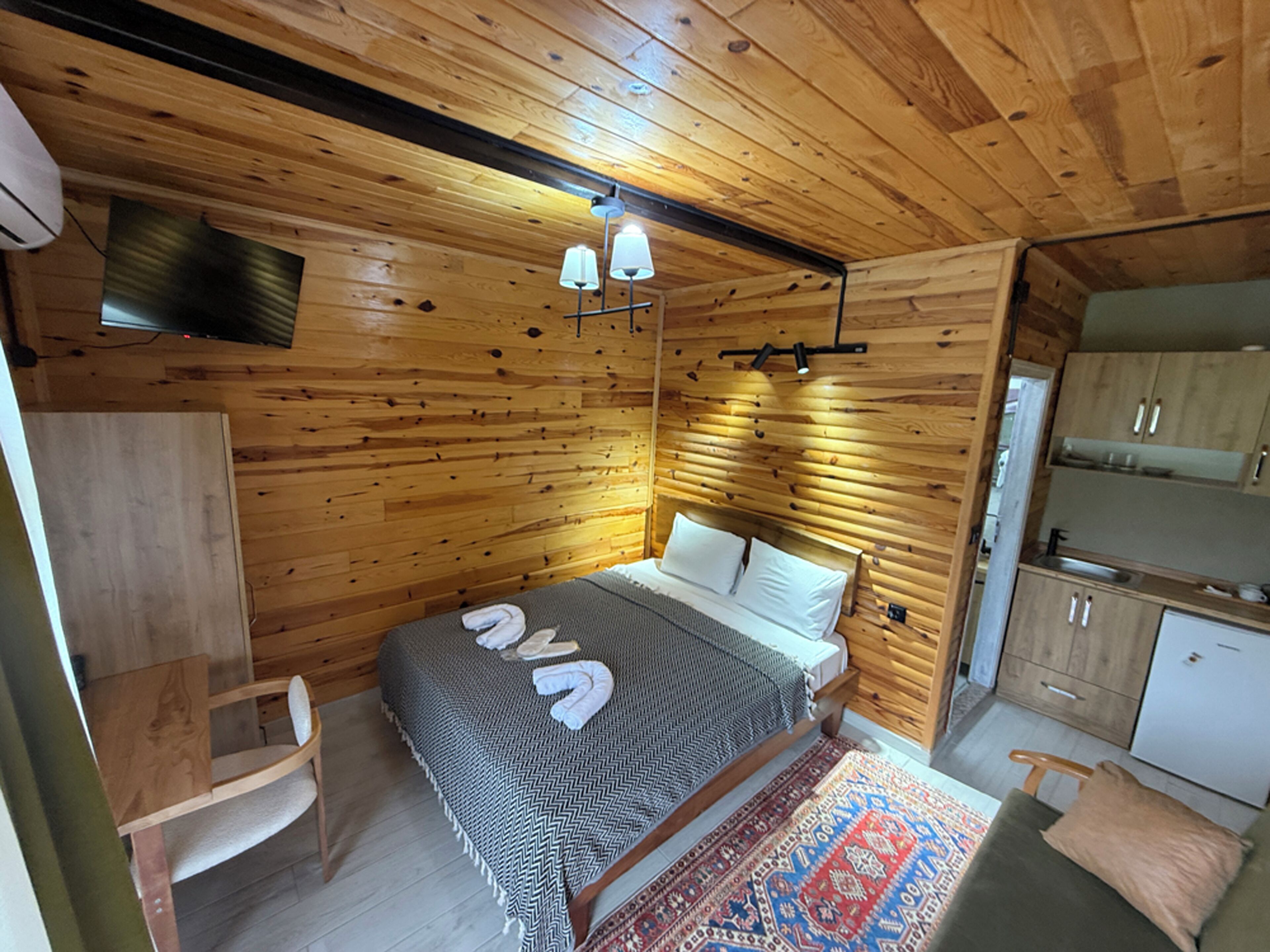 Une pièce aux murs lambrissés, comprenant un lit, un petit bureau, une chaise et un coin cuisine.