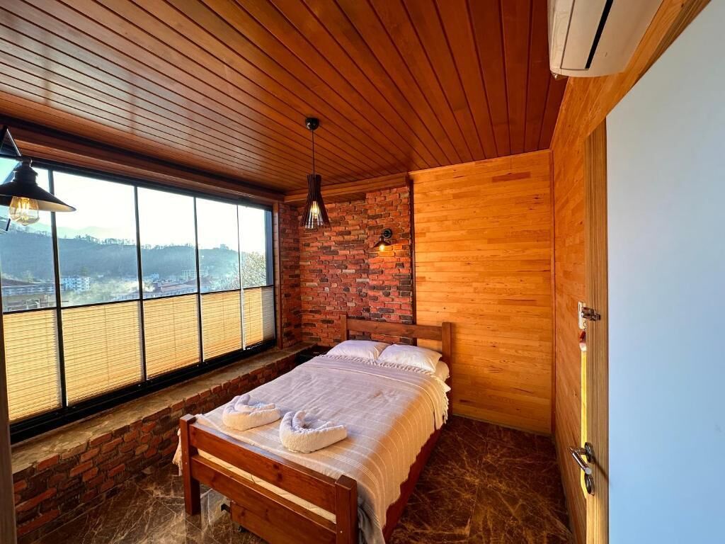 A bedroom with a wooden ceiling, a brick wall, a large window, and a bed with white linens.