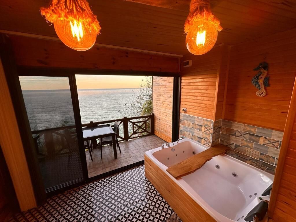 A wooden cabin with a large bathtub, a dining area with a view of the sea, and two hanging light fixtures.