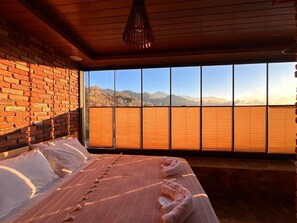 A room with a bed, a brick wall, and a large window with a view of mountains.