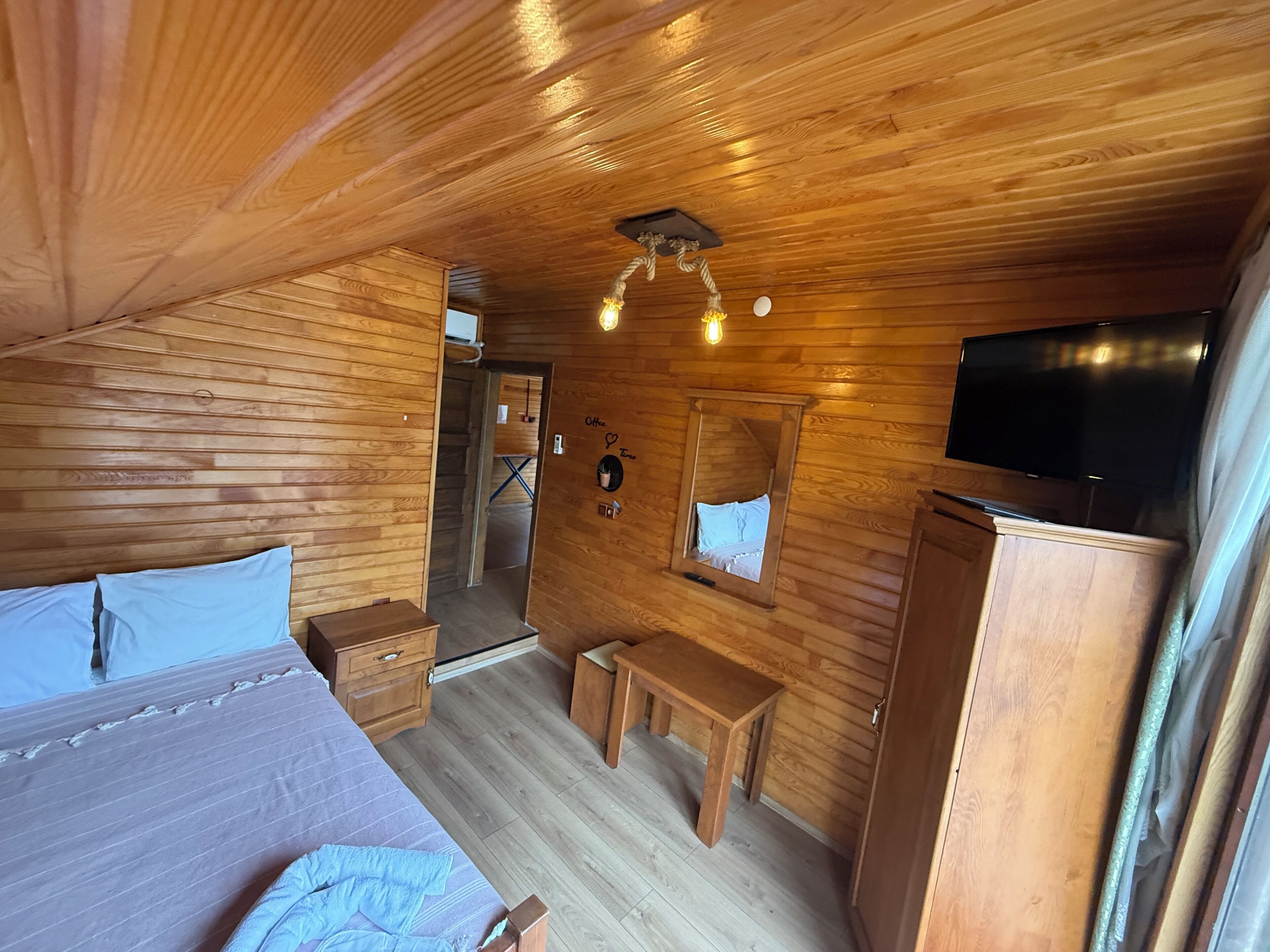 A wooden cabin room with a bed, a desk, a mirror, and a television.
