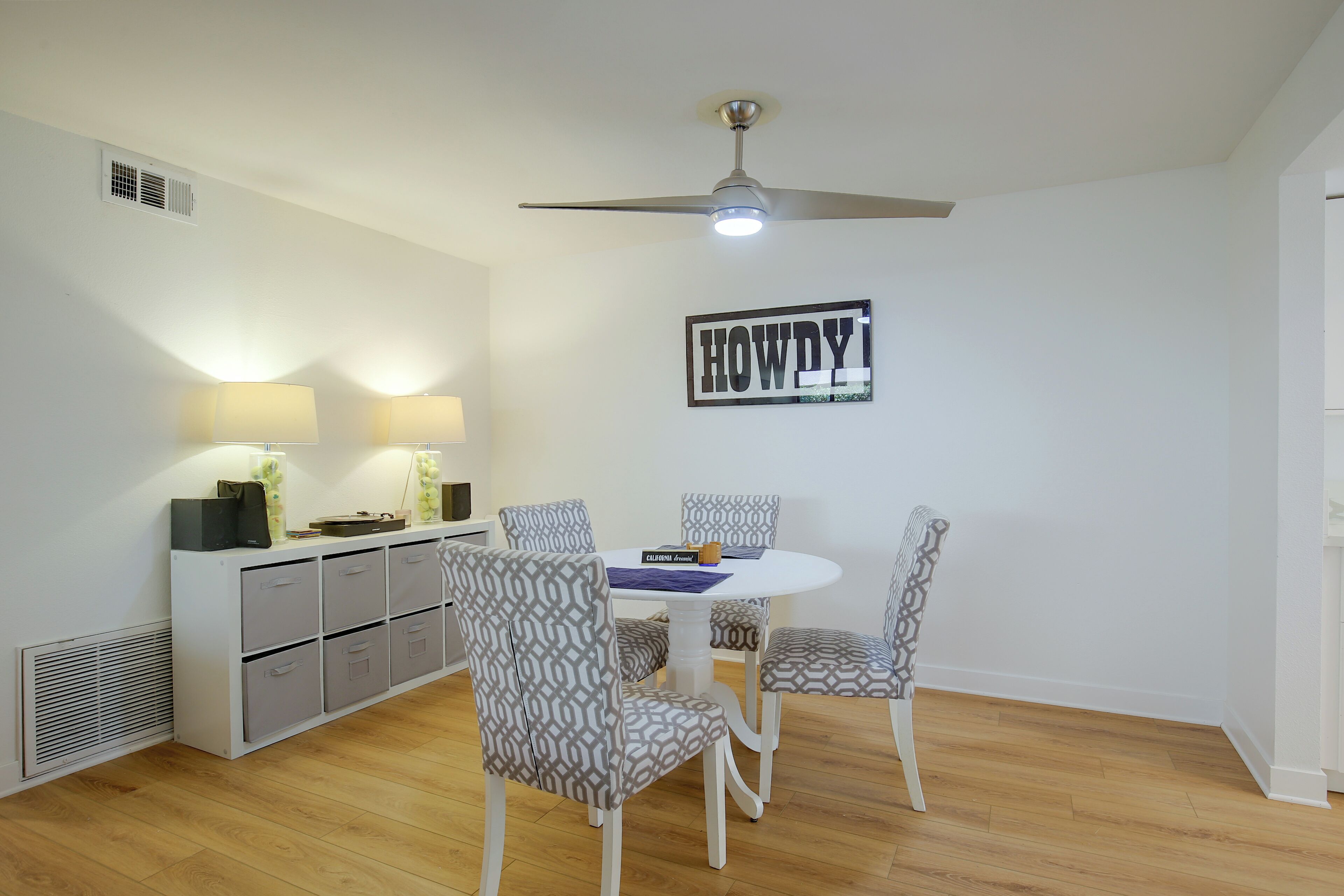 Uma sala de jantar com uma mesa branca e cadeiras estampadas, um ventilador de teto e uma placa fixada na parede.