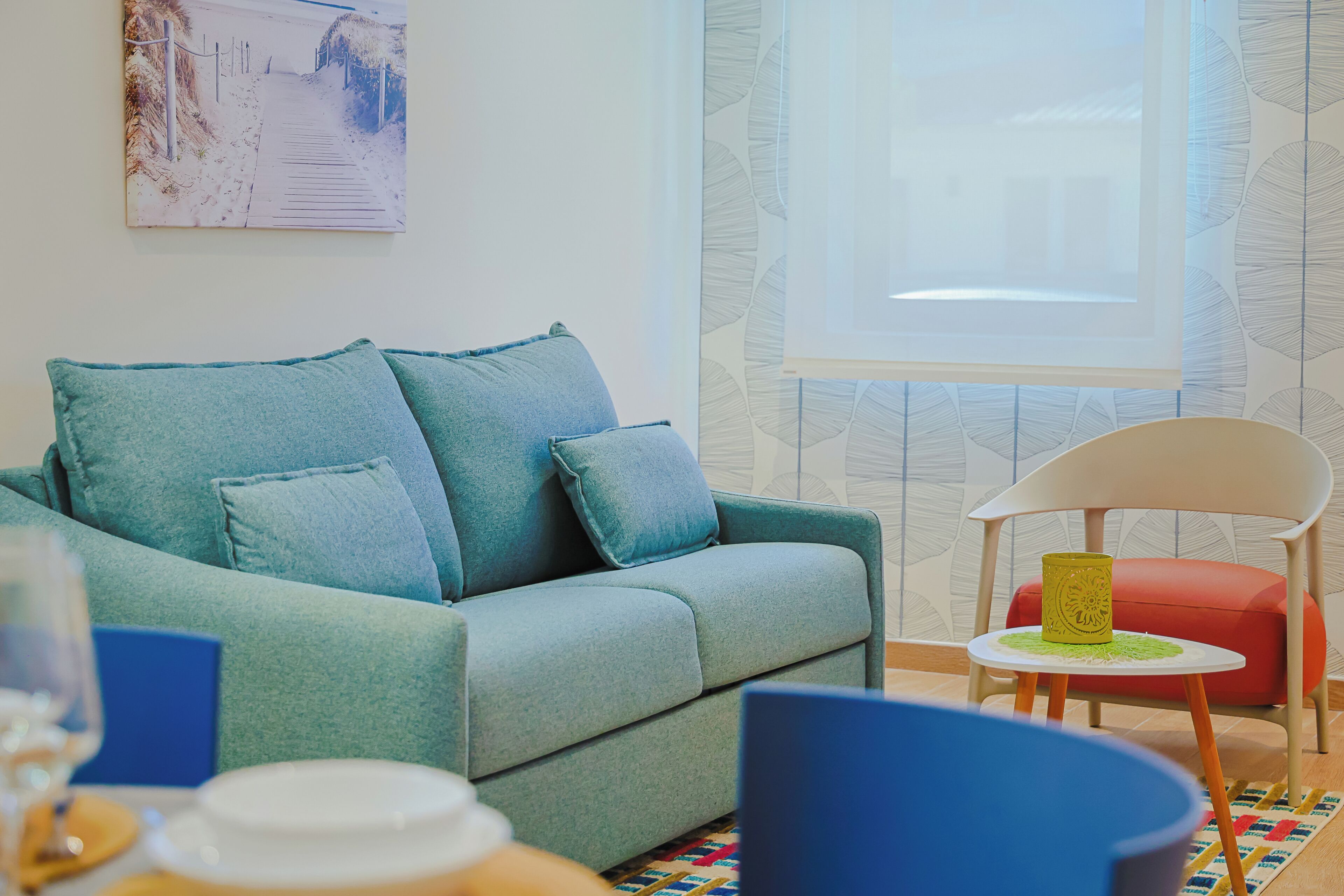 Ein Zimmer mit einem petrolfarbenen Sofa, einem kleinen runden Tisch, einem roten Sessel und einem Fenster mit gemusterten Vorhängen.