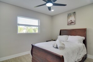 Un dormitorio con cama de madera, ropa de cama blanca, ventilador de techo, ventana con persianas y una obra de arte colgada en la pared.