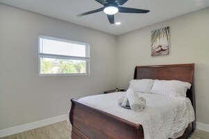 Un dormitorio con cama de madera, ropa de cama blanca, ventilador de techo, ventana con persianas y una obra de arte colgada en la pared.