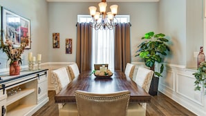 A dining room with a wooden table, six upholstered chairs, a chandelier, a large window with curtains, and a potted plant. - Sun, Fun & Soak: Stunning Myrtle Beach Escape! (Myrtle Beach)