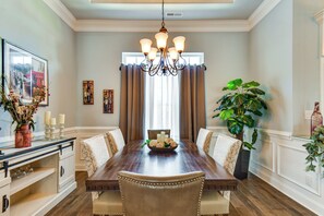 A dining room with a wooden table, six upholstered chairs, a chandelier, a large window with curtains, and a potted plant. - Sun, Fun & Soak: Stunning Myrtle Beach Escape! (Myrtle Beach)