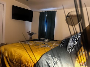 A bedroom with a bed, a flat-screen TV mounted on the wall, a window with curtains, and a vase with dried flowers on a nightstand.