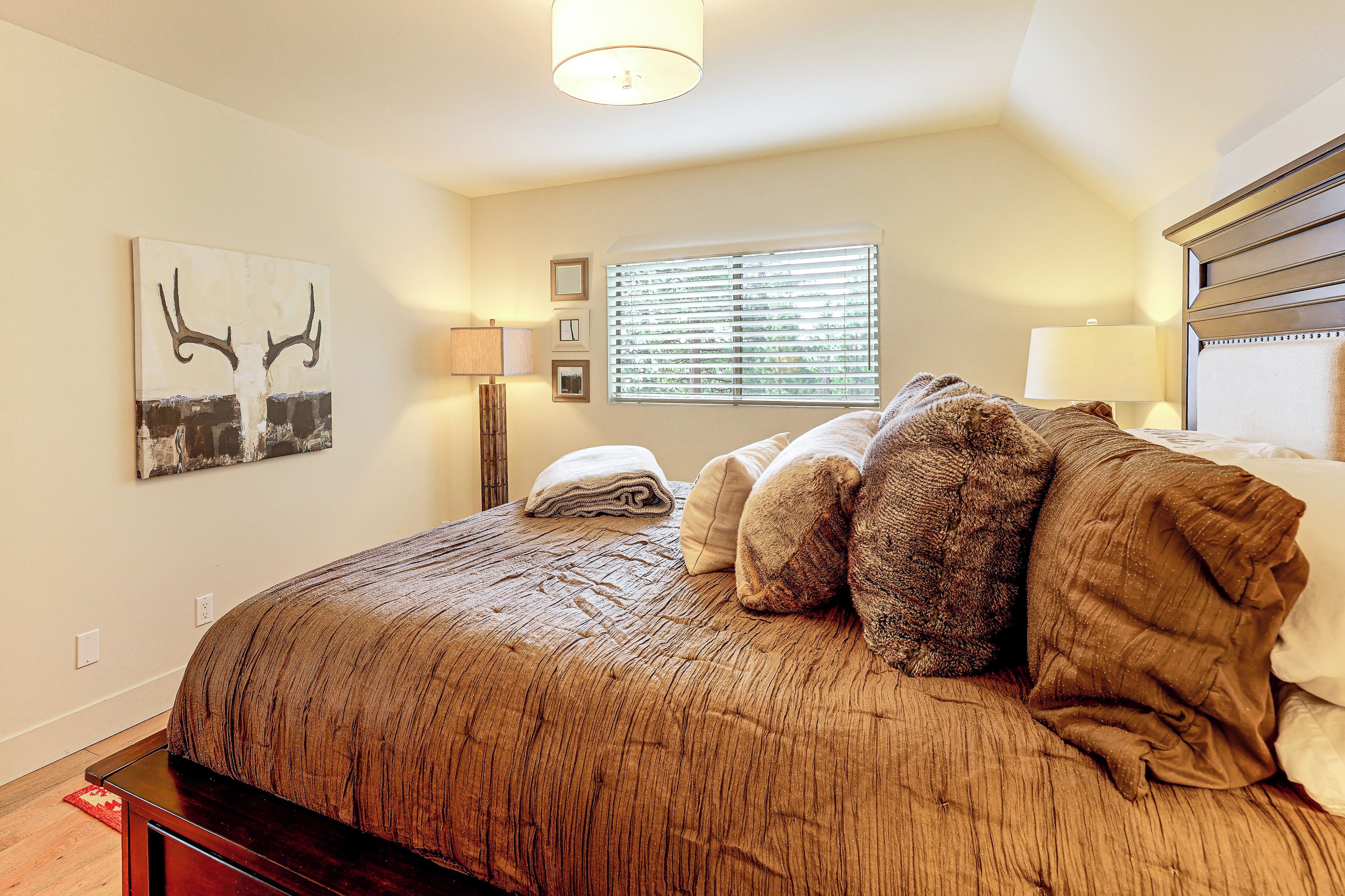 A bedroom with a large bed, a window with blinds, a wall-mounted lamp, and a painting.