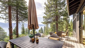 Una terraza de madera con mobiliario exterior, una mesa y una sombrilla, con vistas a un lago y un bosque.