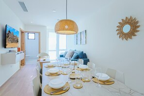 A modern dining area with a long table set for a meal, a television on the wall, and a sun-shaped mirror decoration. - Bonsai 3BR Retreat Grand 207 (Noord)