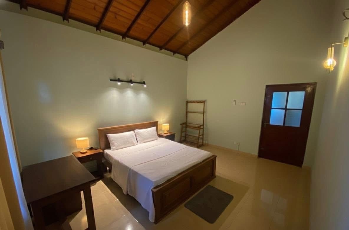 A bedroom with a wooden bed, bedside lamps, a nightstand, a small table, a ladder, and a door with a window.