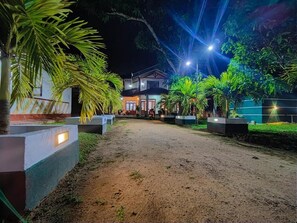A pathway leading to a building with a porch, surrounded by palm trees and illuminated by overhead lights.