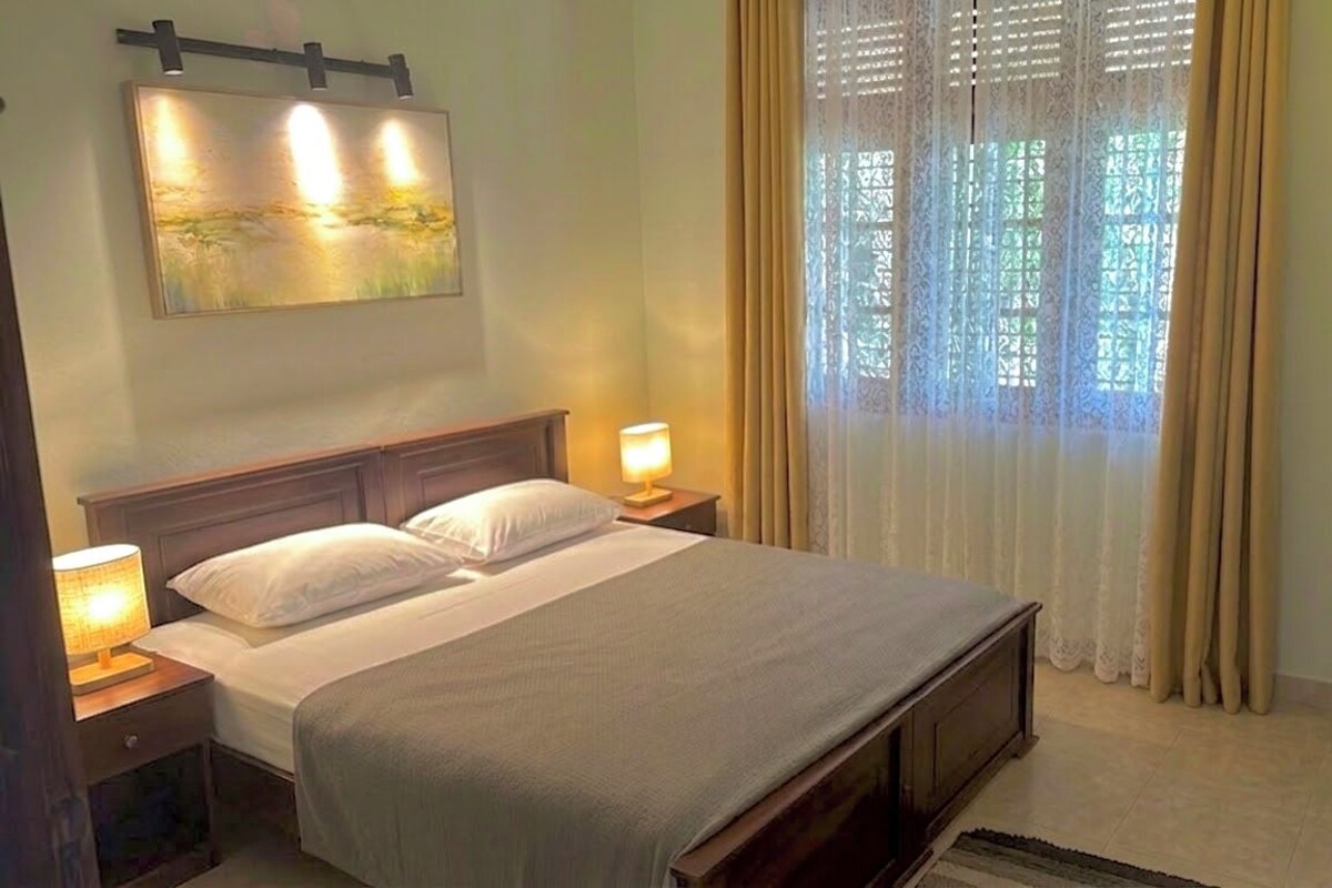 A hotel room with a bed, two bedside tables with lamps, a painting on the wall, and a window with curtains.