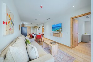 A modern living room with a sofa, dining table, and chairs, a flat-screen TV mounted on the wall, and a view of the ocean through a window. - Bonsai 3BR Retreat Grand 307 (Noord)