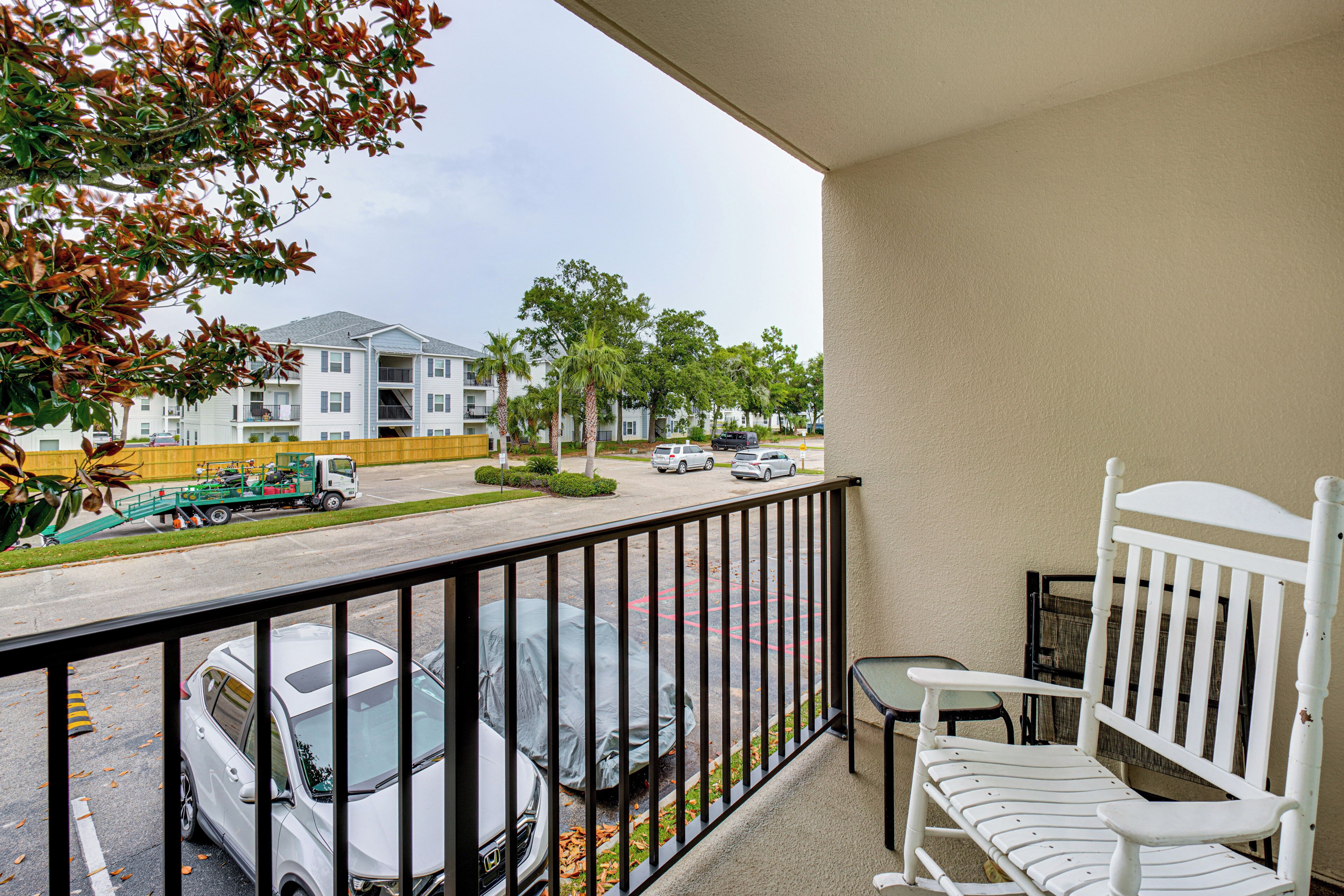 A balcony with a white chair and a small table, overlooking a parking lot with several cars and a green truck.