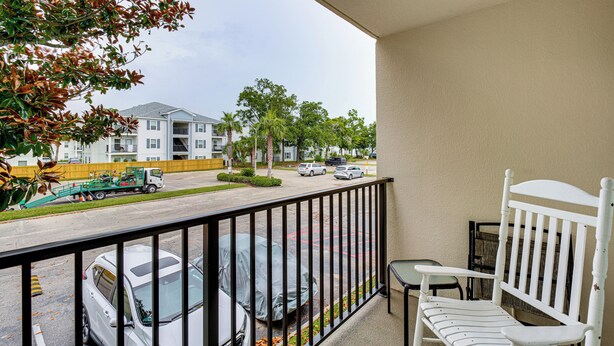 Ein Balkon mit weißem Stuhl und kleinem Tisch, der auf einen Parkplatz mit mehreren Autos und einem grünen Lastwagen blickt.