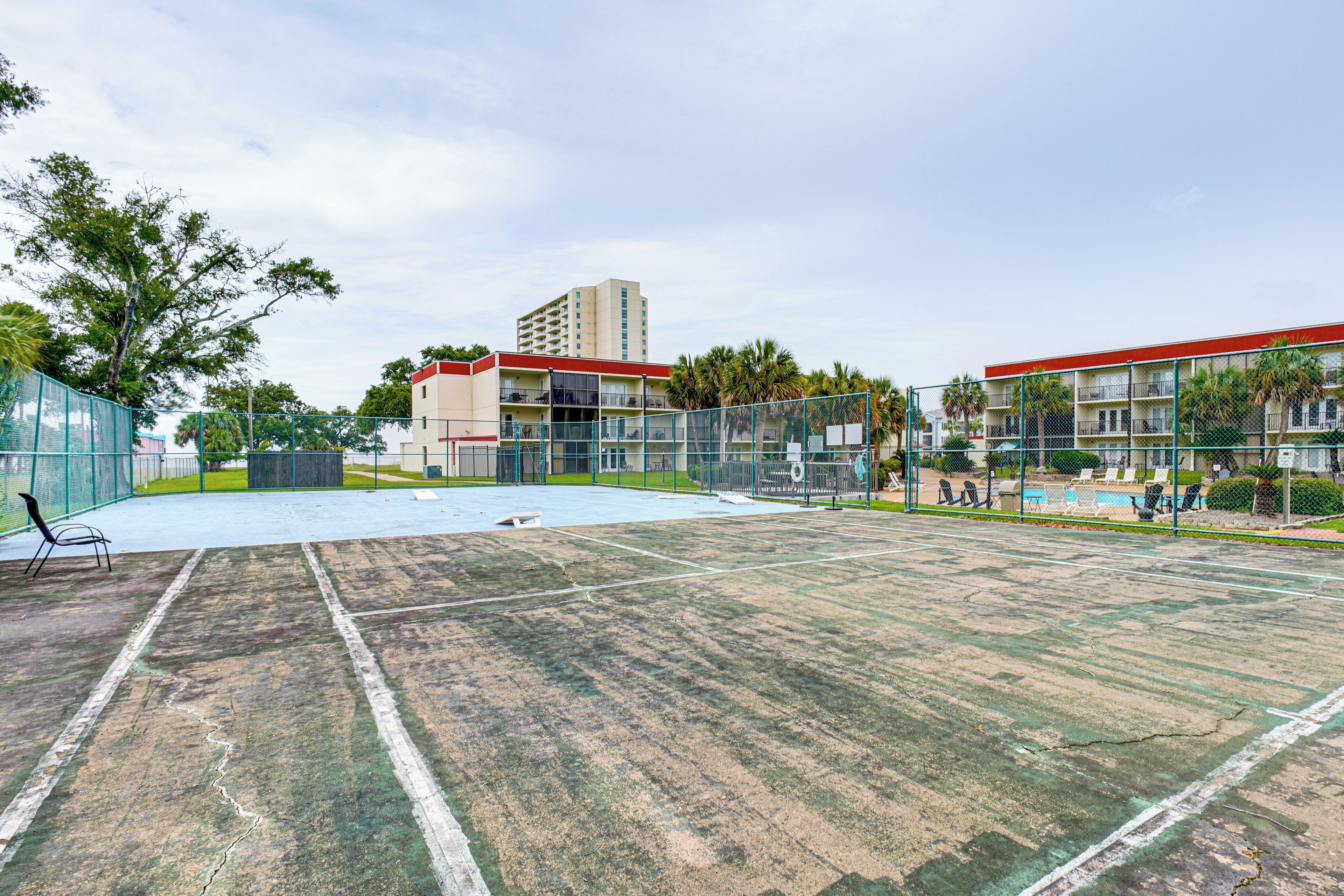 Una cancha de tenis con red, rodeada por una valla, junto a un edificio con varios balcones.