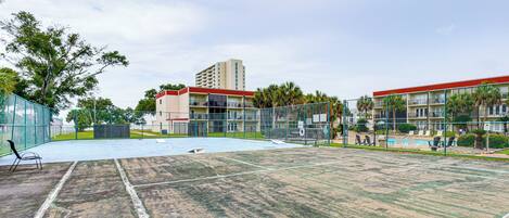 Una cancha de tenis con red, rodeada por una valla, junto a un edificio con varios balcones.