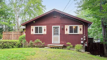 A single-story house with a red exterior, a front door, two windows with shutters, and a small porch.