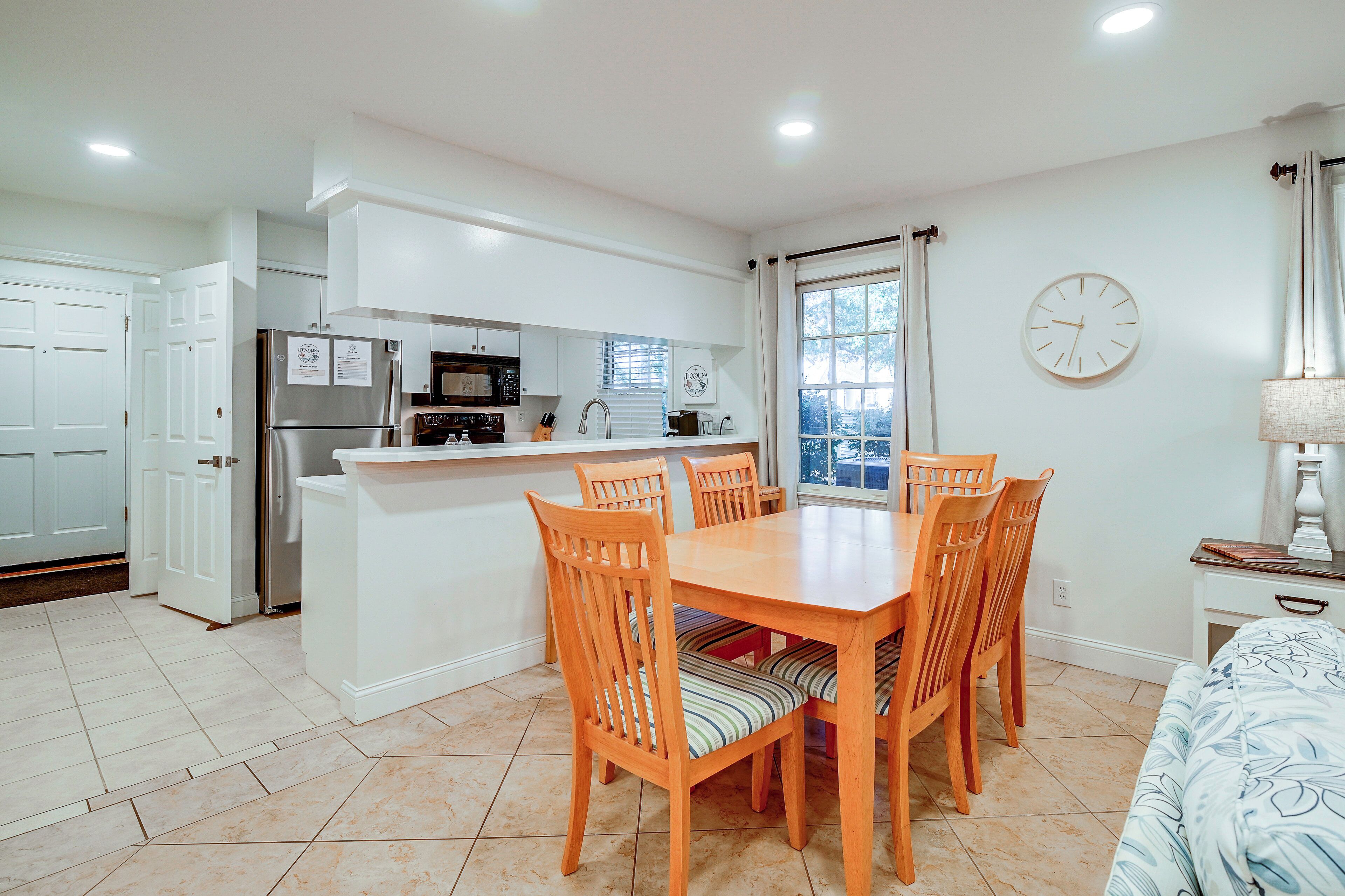 A compact kitchen with modern appliances, a dining area with wooden chairs, and a large window with blinds.