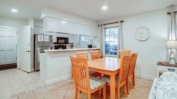 A compact kitchen with modern appliances, a dining area with wooden chairs, and a large window with blinds.