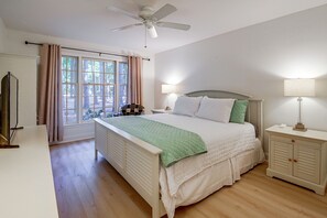 Quarto com cama, mesa de cabeceira, luminária, ventilador de teto e janela com cortinas.