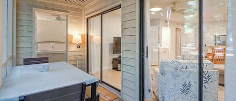 A room with a bed, a table, and a chair, with a view of another room through a glass door.