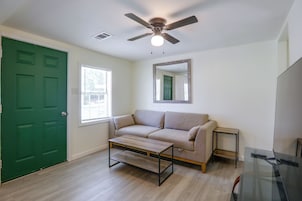 Una habitación con puerta verde, sofá color beige, una mesita pequeña, ventilador de techo y ventana con persianas.