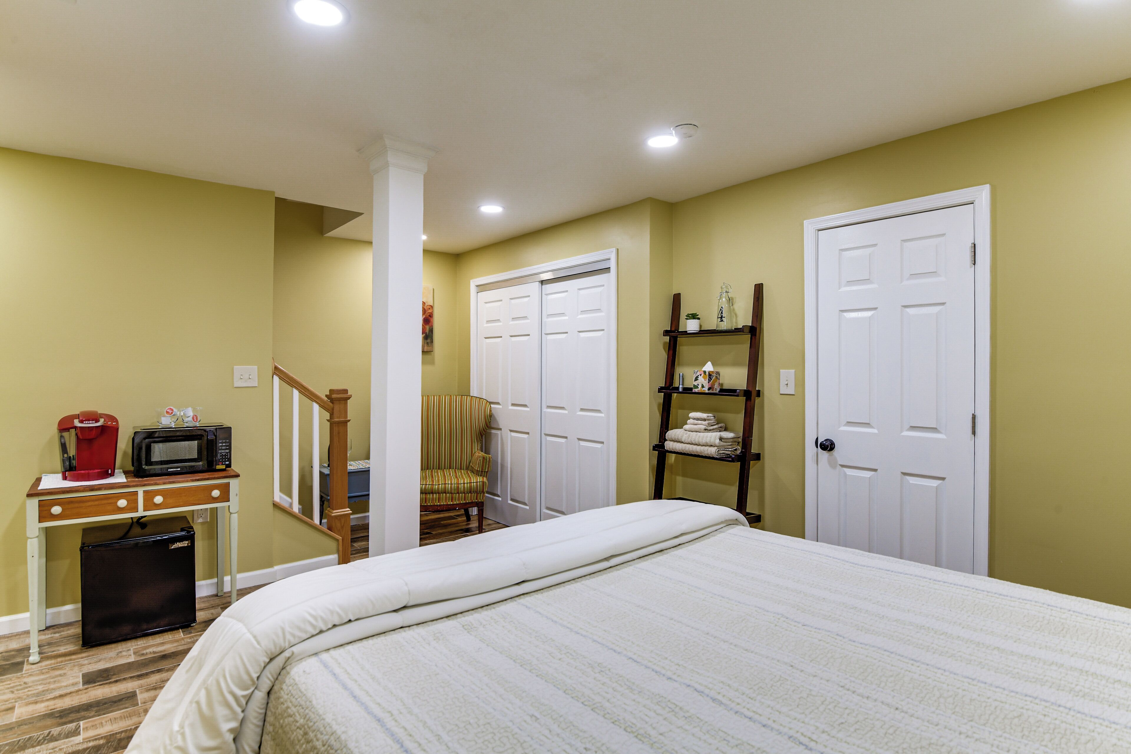 A bedroom with a bed, a desk with a coffee maker, a chair, a shelf with towels, and a door.