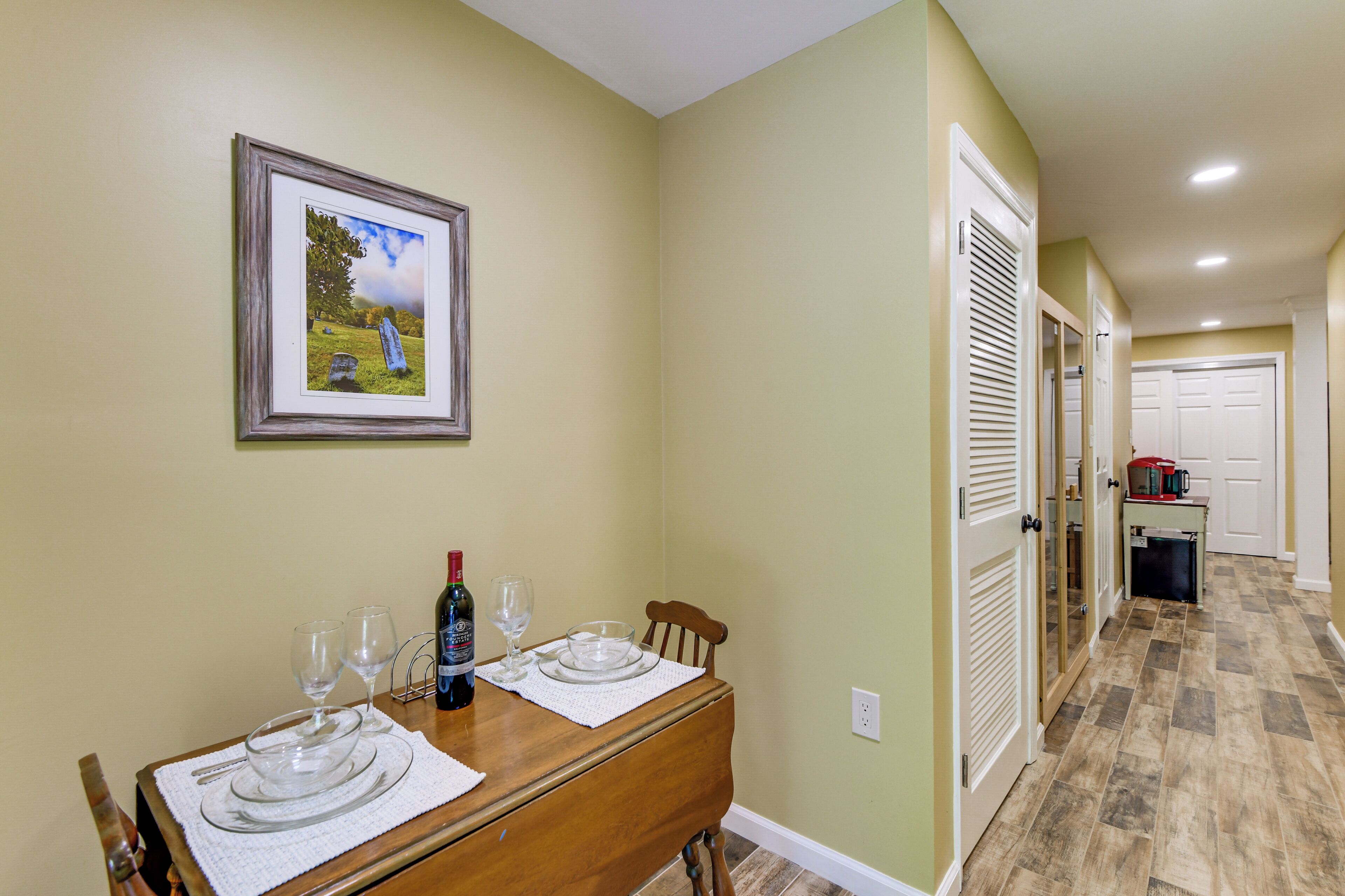 A dining area with a table set for two, a framed picture on the wall, and a kitchen visible in the background.
